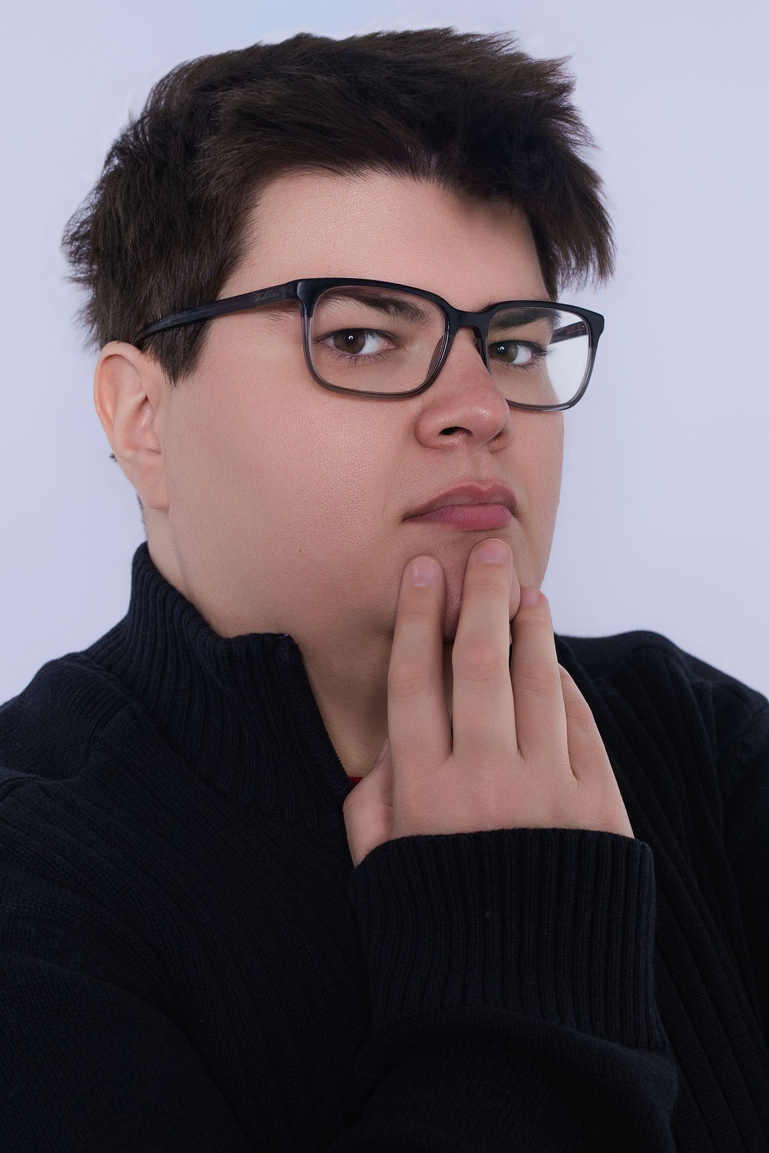 Young person wearing glasses and black turtleneck poses thoughtfully with hand on chin.