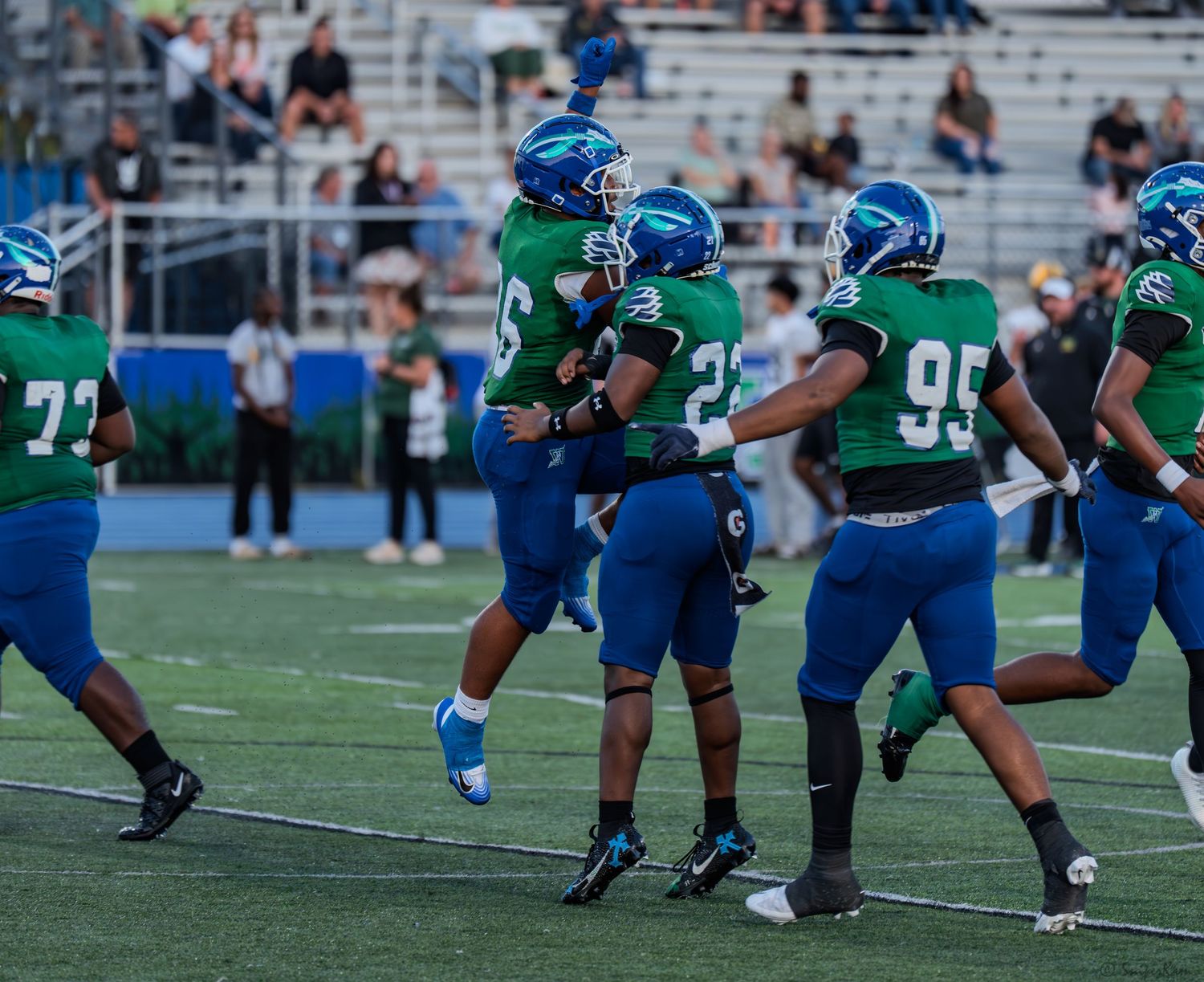 High school football players in blue and green uniforms celebrate with a chest bump during a night game.