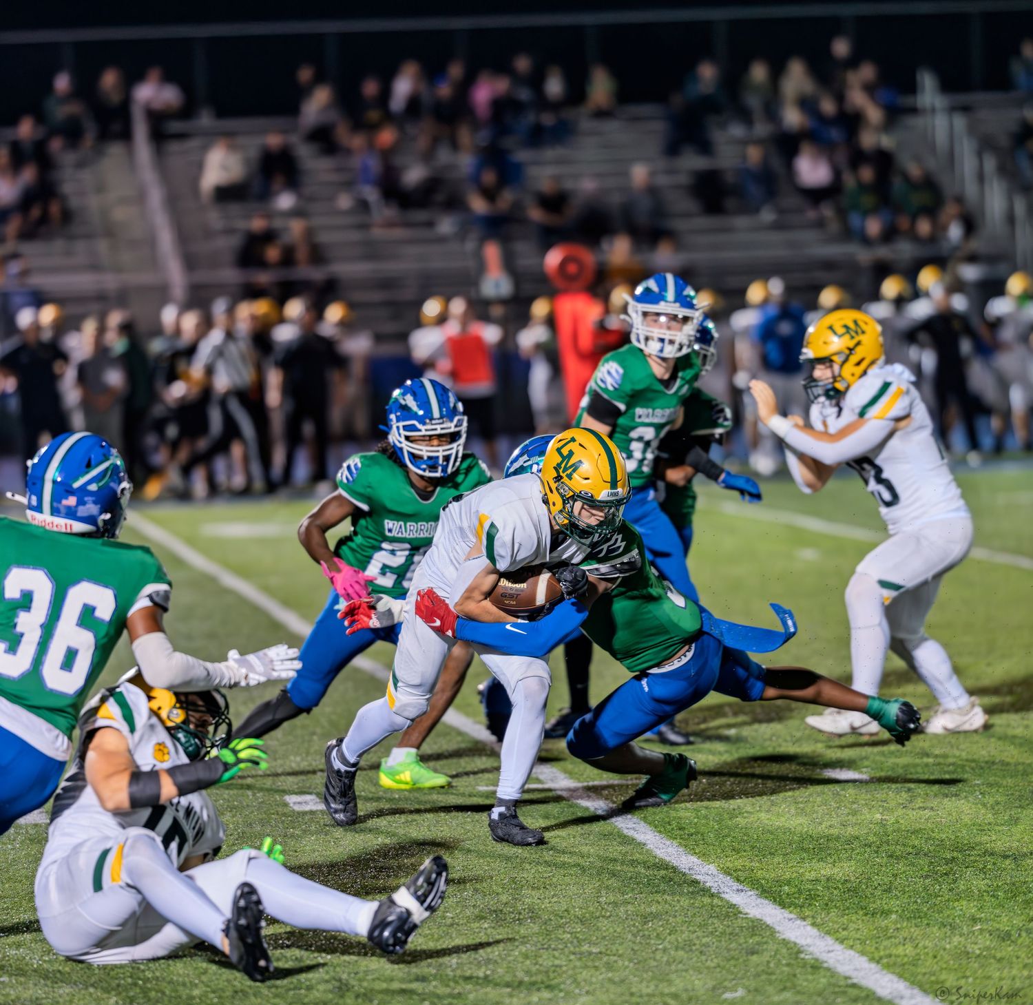 High school football player in gold uniform gets tackled by defenders in green and blue during a night game.
