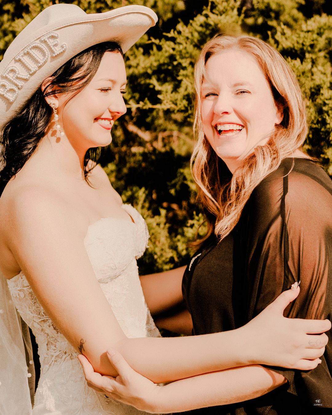 Laughing bride wearing BRIDE cowboy hat embracing smiling woman in black outfit outdoors.