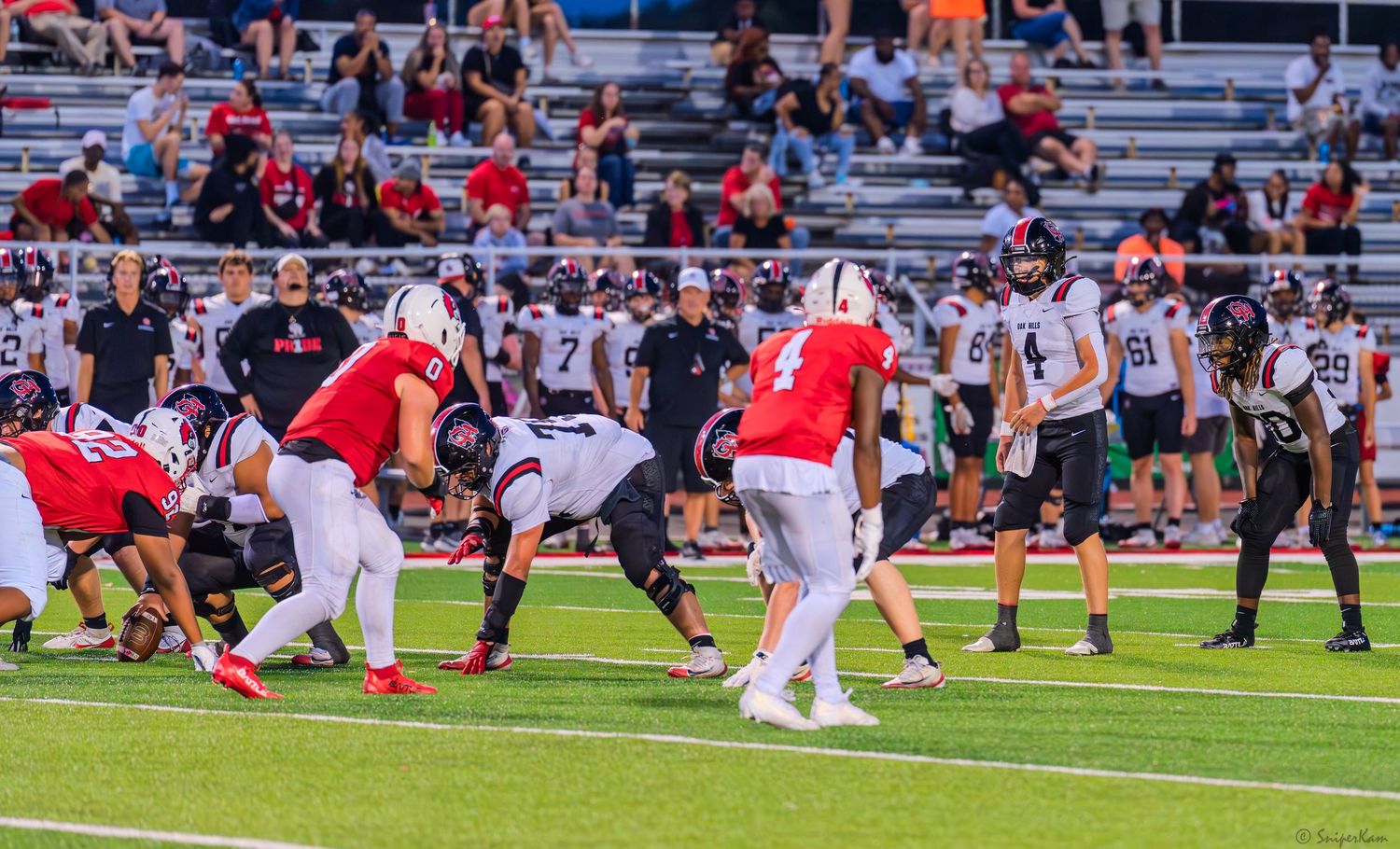High school football players compete during a night game on a red turf field with fans watching.