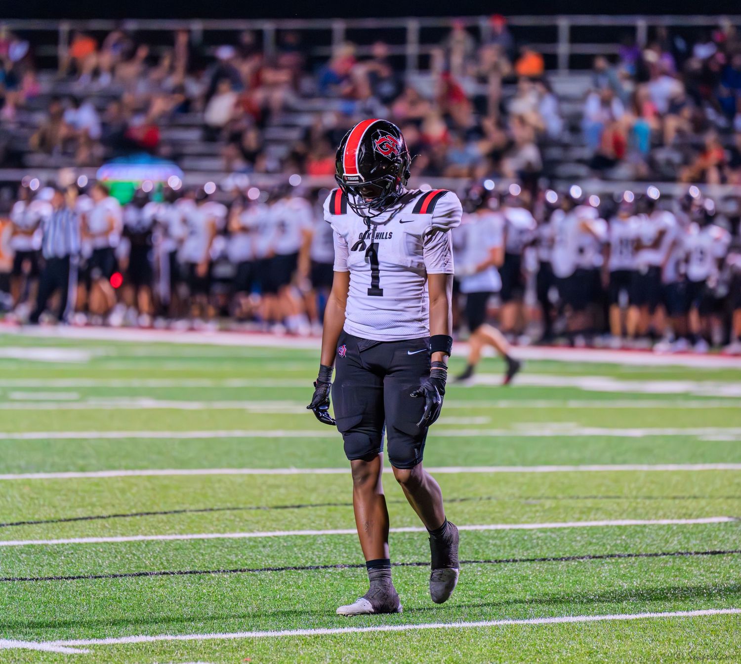 A football player wearing #1 in red and white uniform stands alone on the field during a high school night game.