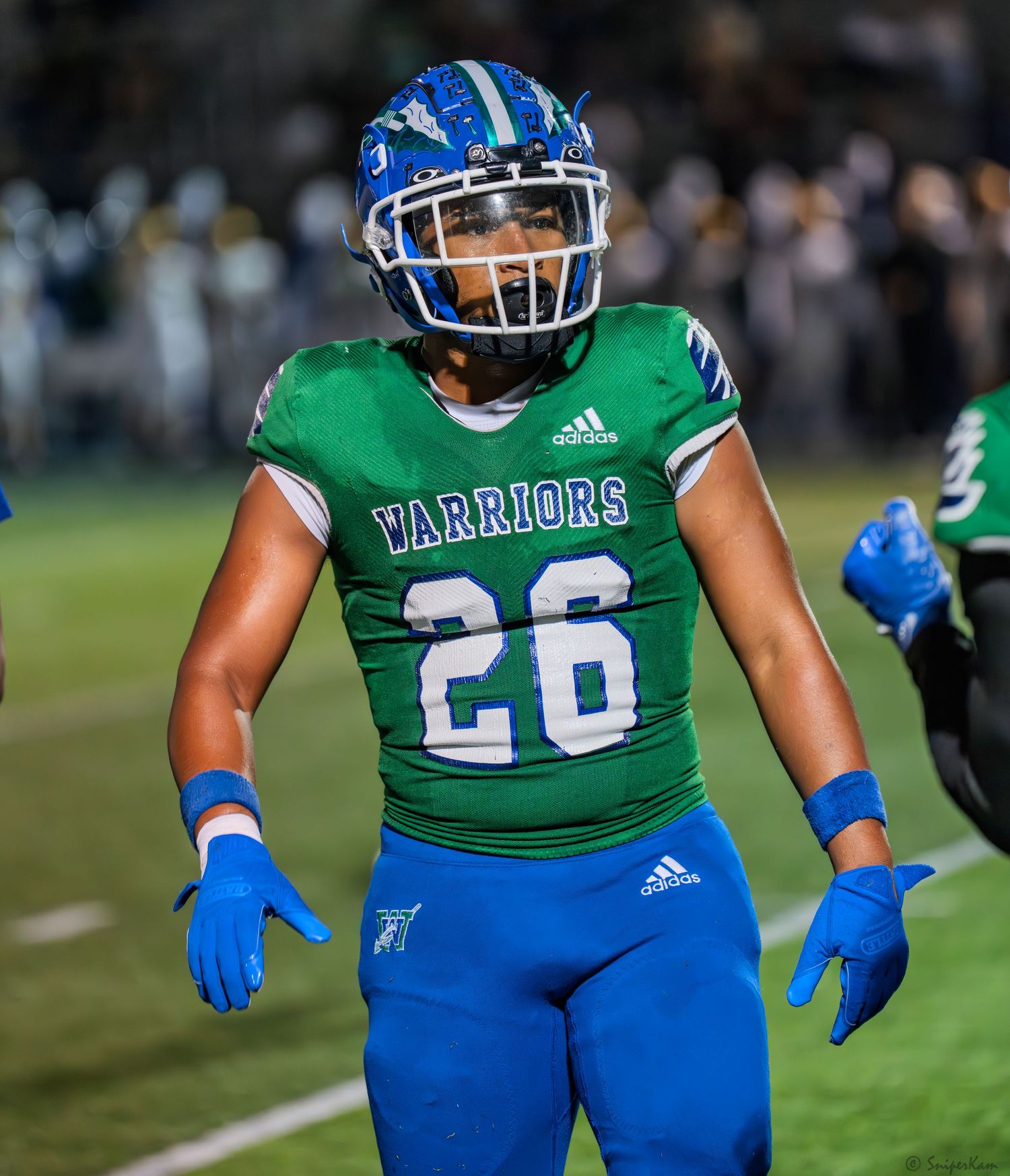 Warriors football player #26 in green Adidas uniform stands on field during high school game.