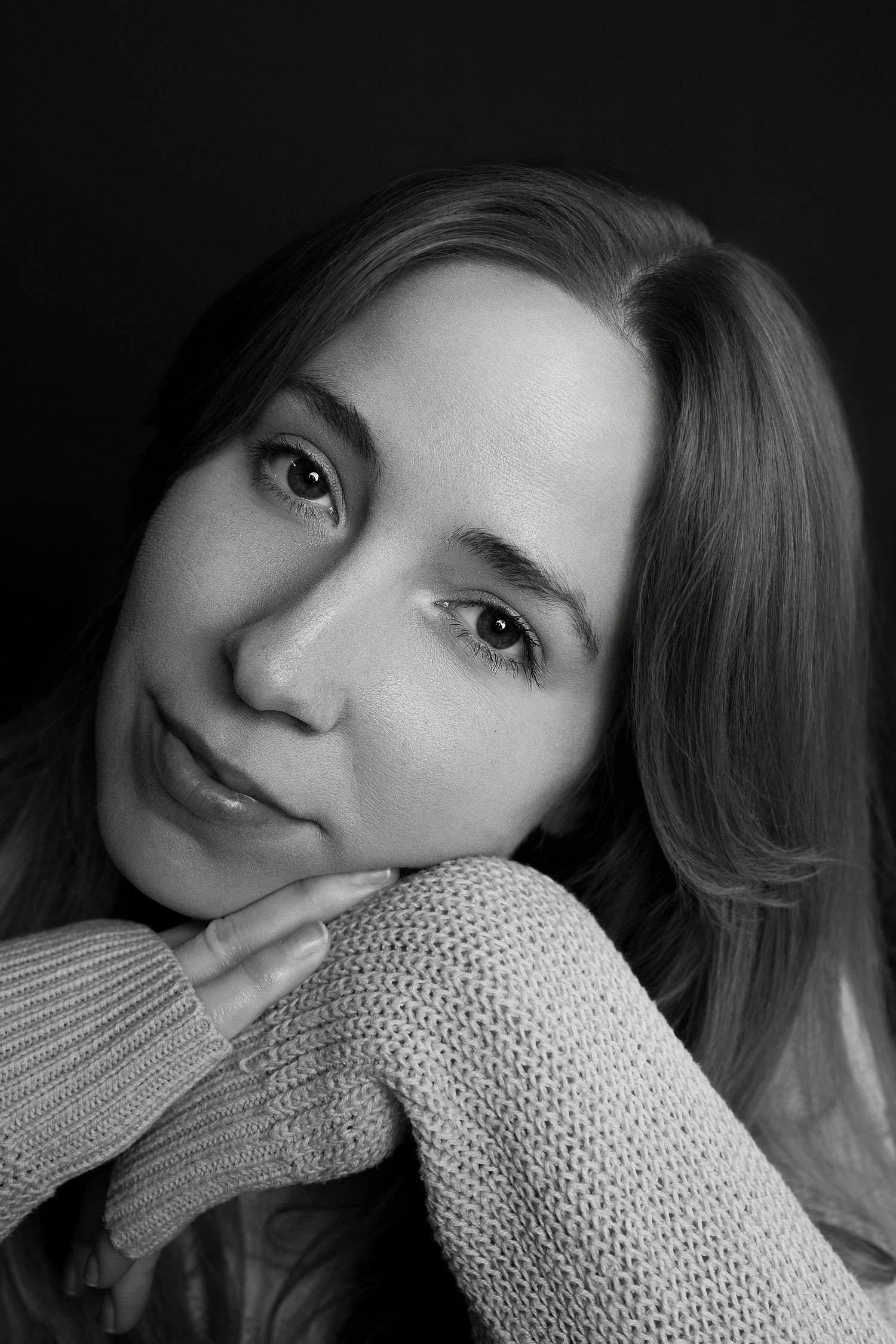 Black and white close-up portrait of young woman resting chin on hands wearing knit sweater.