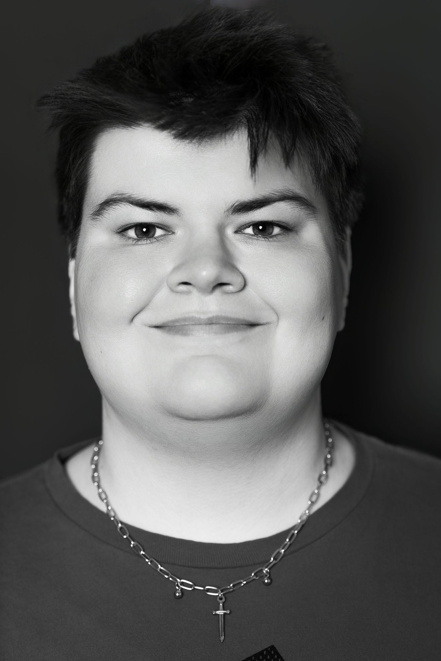Black and white portrait of a smiling young person wearing a silver chain necklace with a cross pendant.