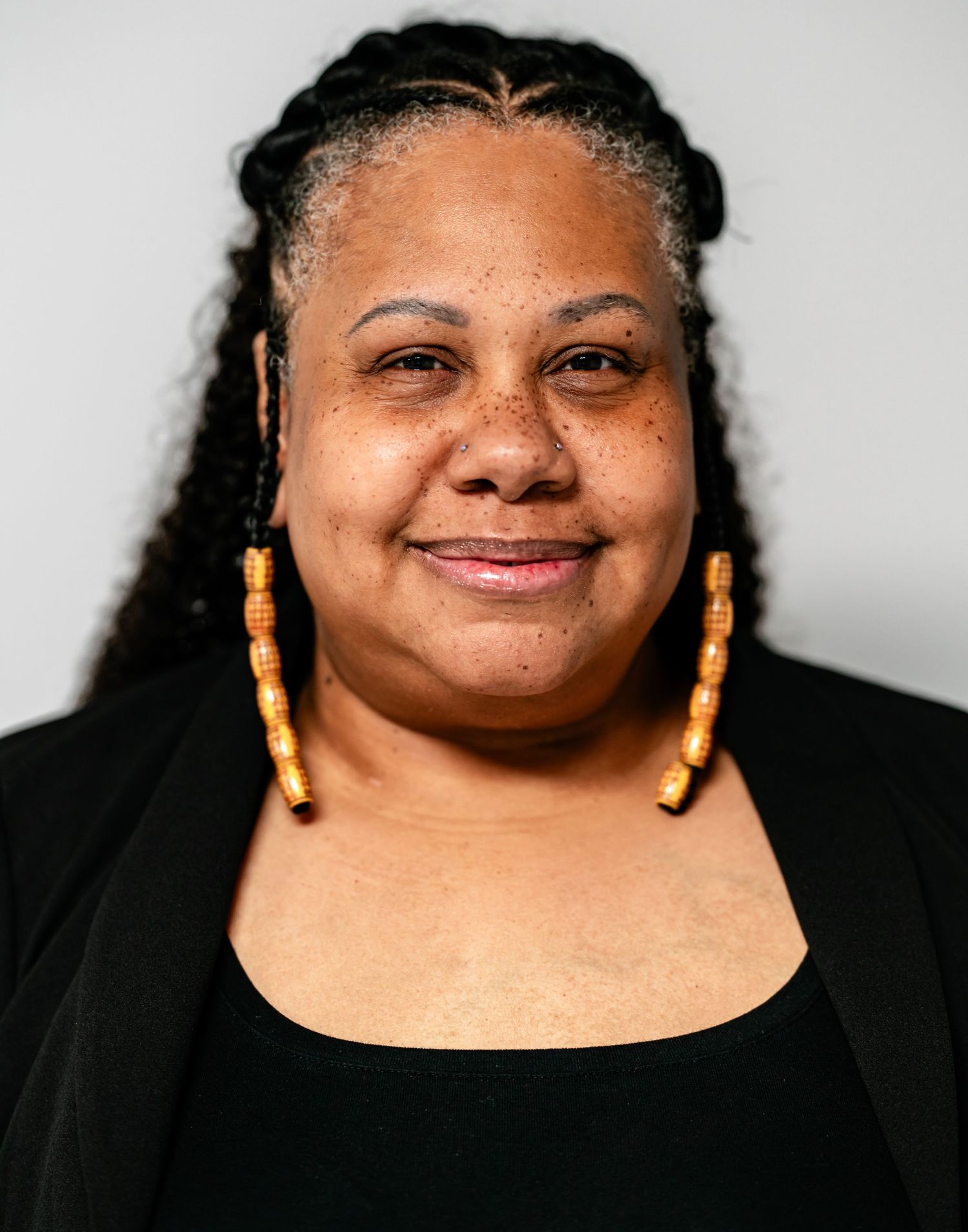 A smiling Black woman with long braids and orange beaded earrings poses in a black outfit against a light background.