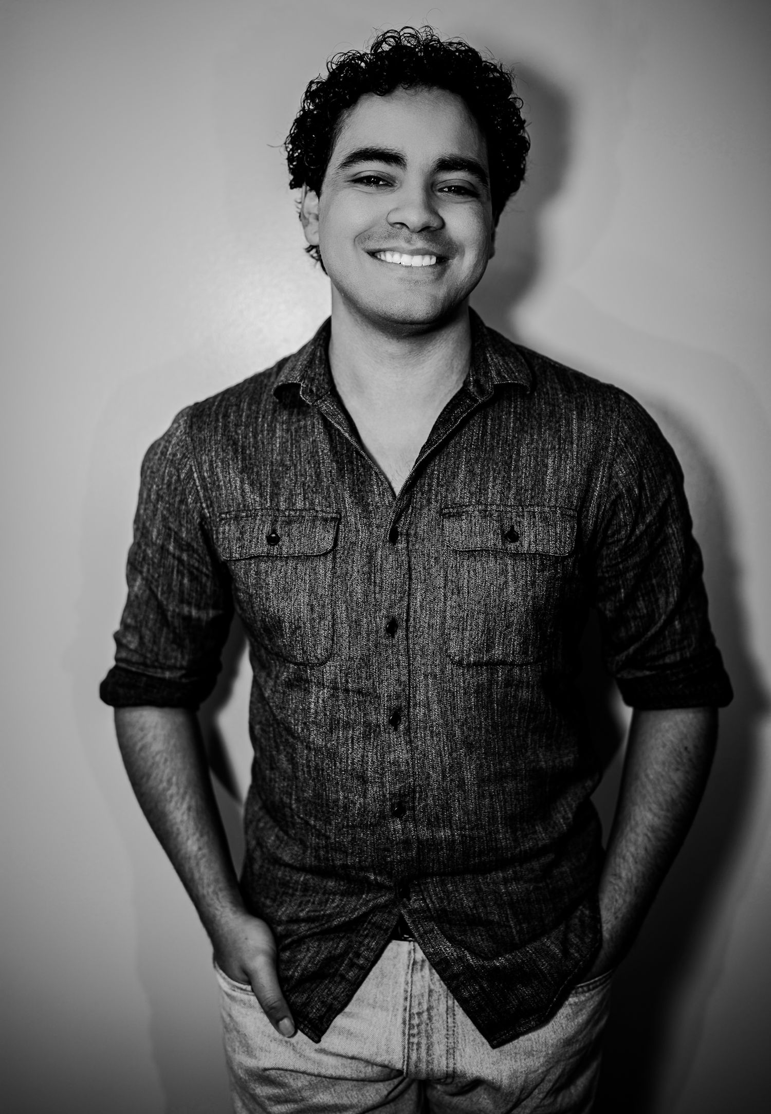 Black and white photo of smiling young man in button-up shirt with hands in pockets.