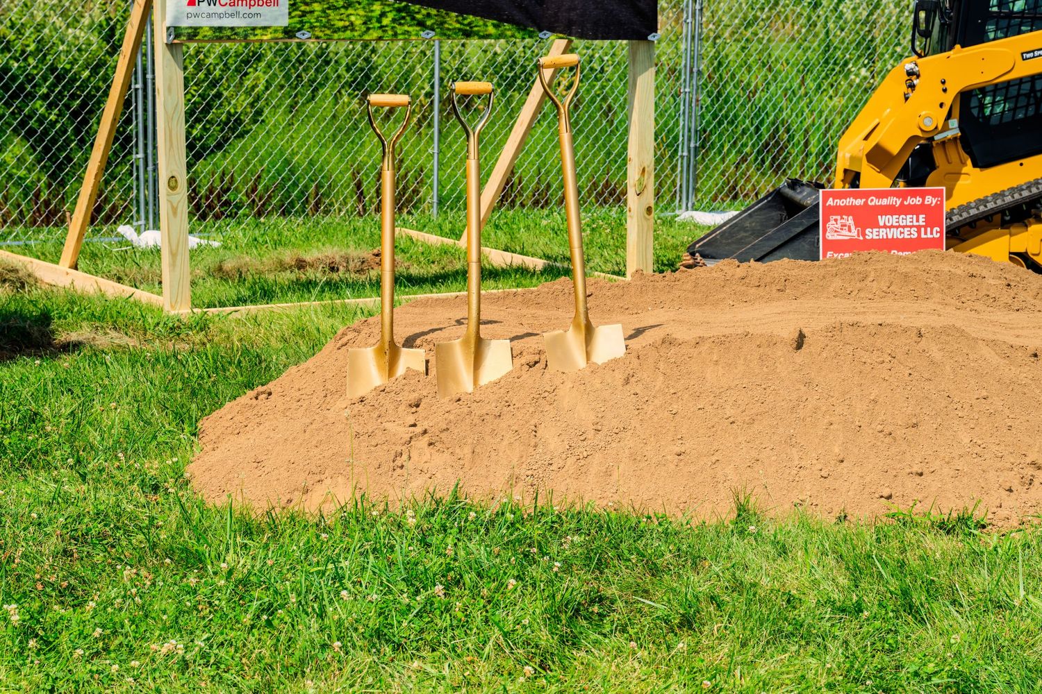Golden ceremonial shovels standing in dirt mound at COPCU groundbreaking site with yellow excavator nearby.