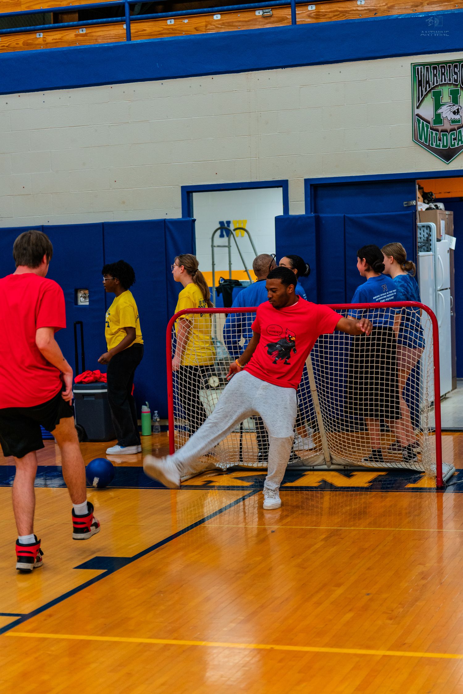 Students playing indoor floor hockey in a school gymnasium, with a player in red defending the goal net.