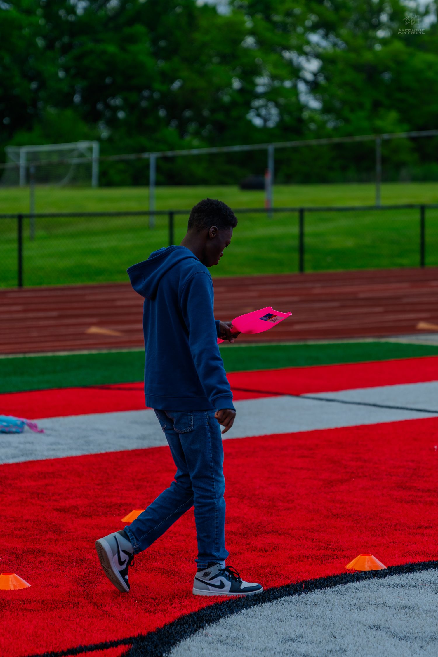 Young man in blue hoodie and jeans walking on red turf field holding a pink clipboard.