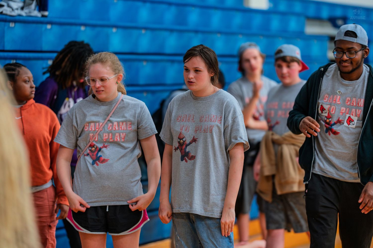 Students wearing matching gray camp t-shirts walk together inside a school gymnasium during a group activity.