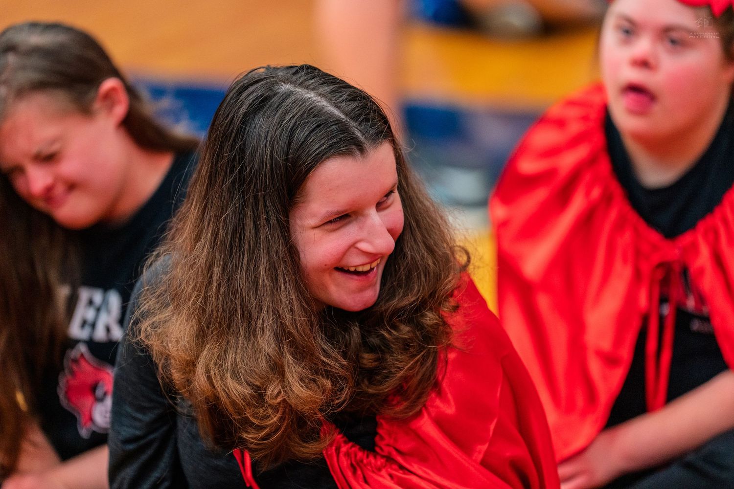 Young woman laughing joyfully at a special needs sports event, wearing red costume, with peers in background.