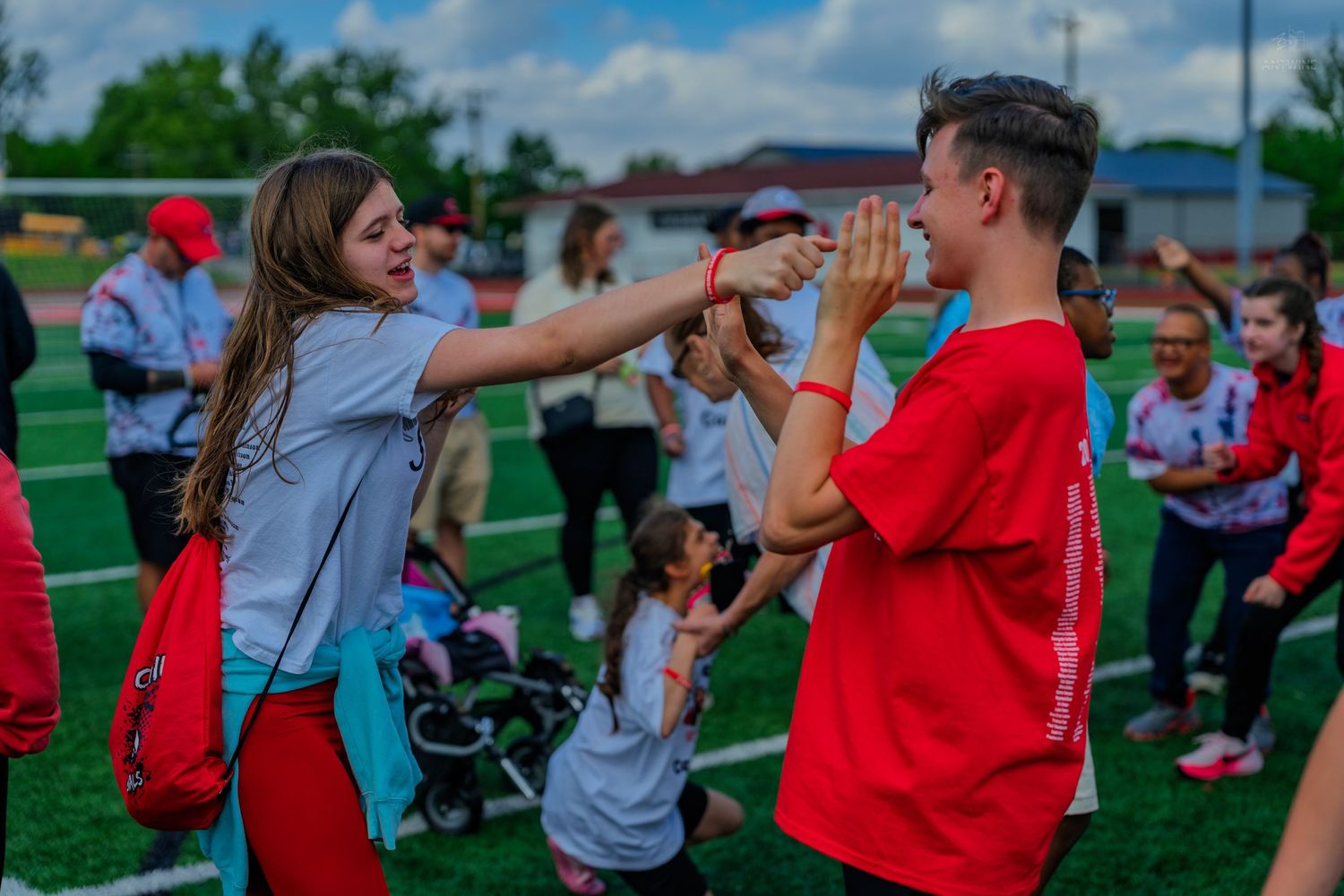 Two teens in red shirts high-five on a green sports field during an outdoor event with other participants nearby.