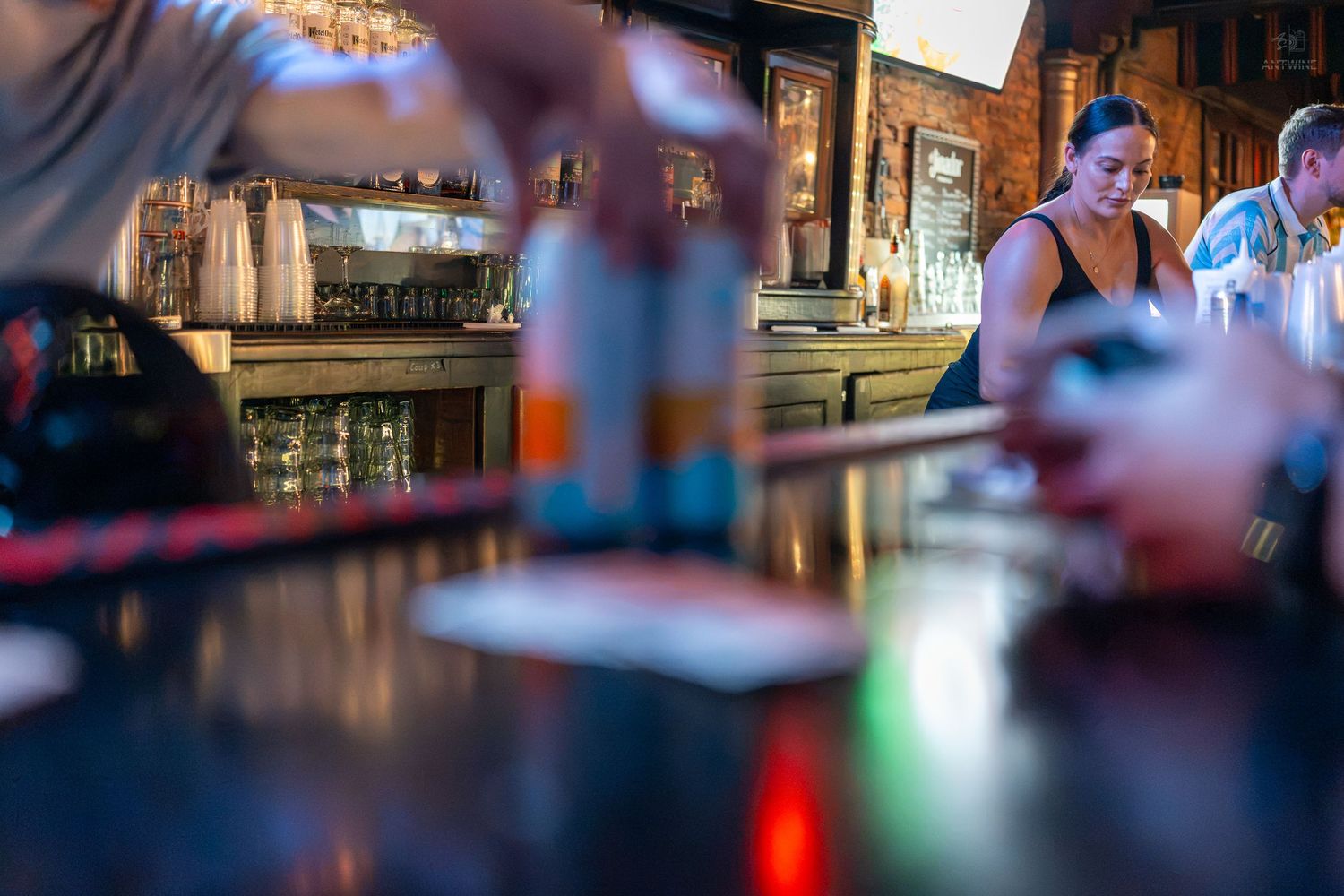 Woman bartender working behind a bar counter in a dimly lit pub with bottles and shelves in background.