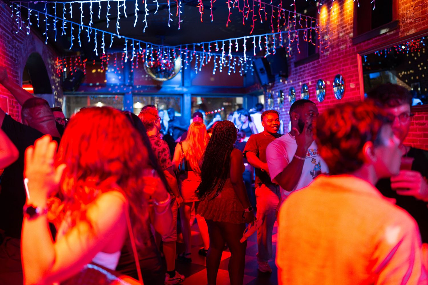 Vibrant nightclub scene with people dancing under colorful red and blue lights with string lights overhead.