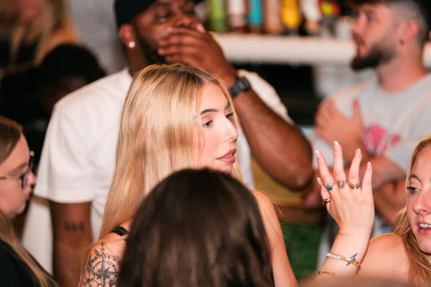 Blonde woman gesturing with ringed hands while socializing at a lively bar event.