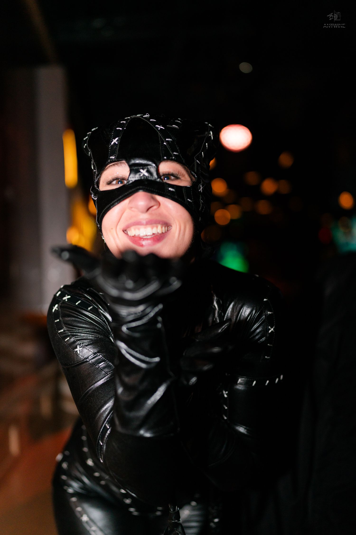 Woman in shiny black Catwoman costume and mask smiles and leans toward camera at night.