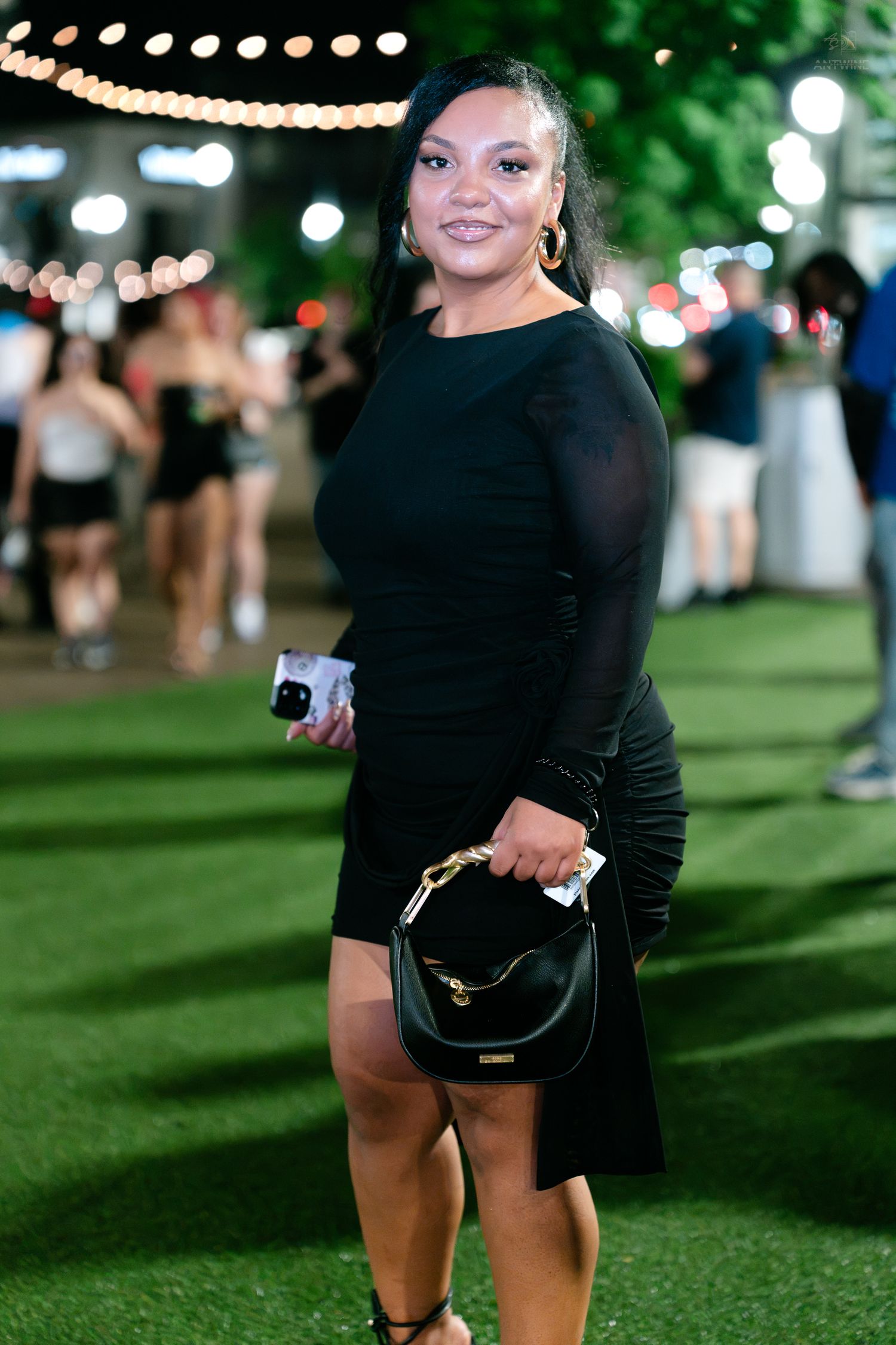 Smiling woman in black long sleeve mini dress holding handbag at outdoor night event.