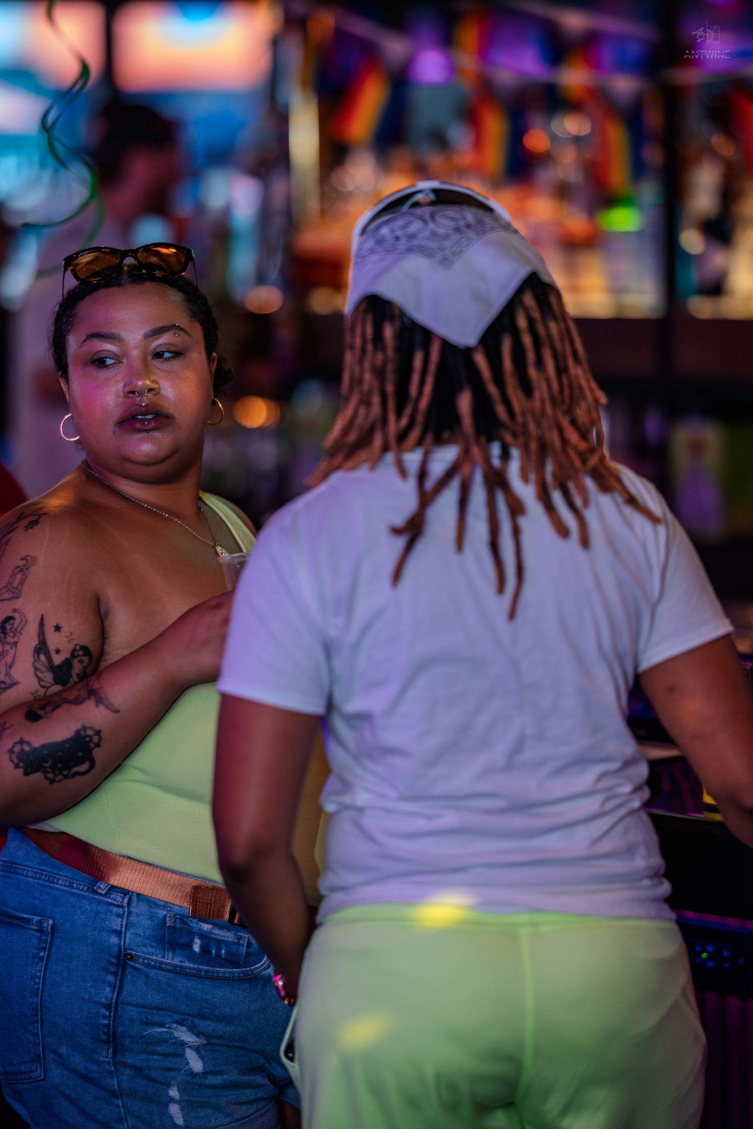 Two women interact near a neon-lit bar, one with dreadlocks in a white shirt.