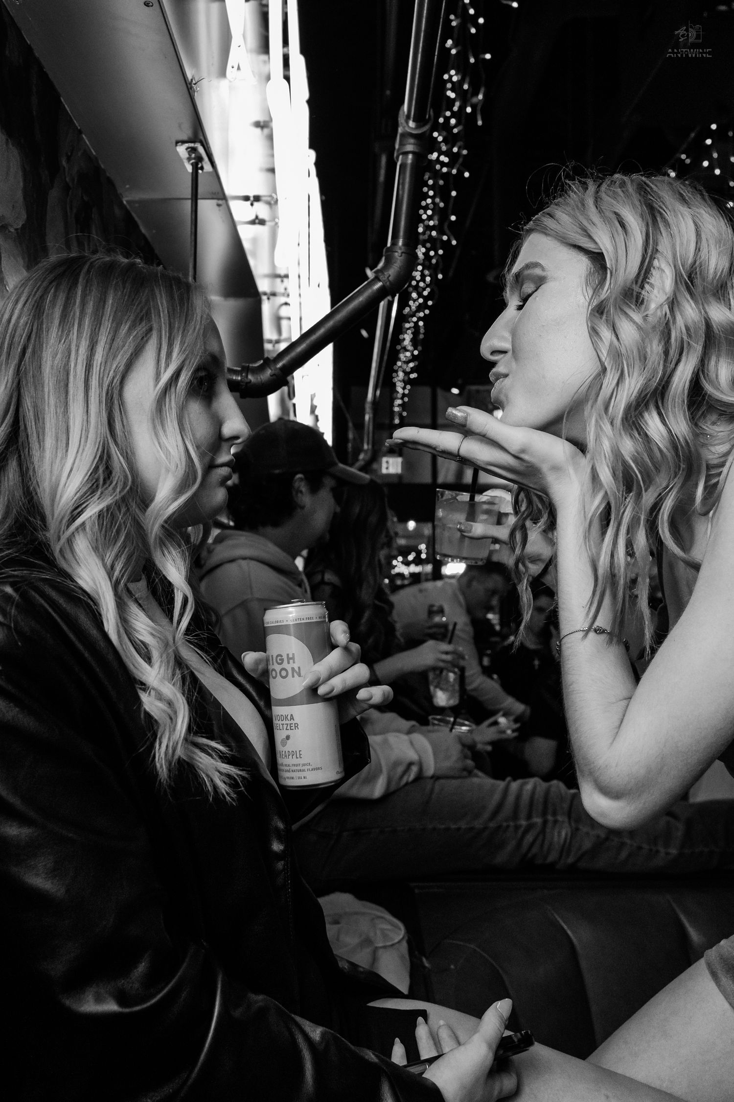 Two blonde women share a lighthearted moment outside a bar, one holding a canned drink at night.