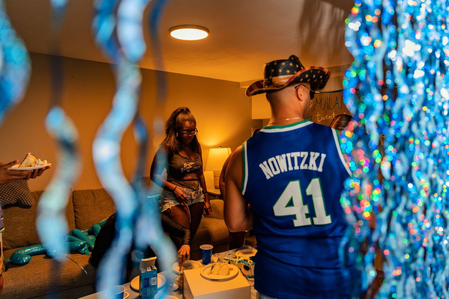 Party guests socializing in a blue-decorated room, with a man wearing a Nowitzki 41 basketball jersey.