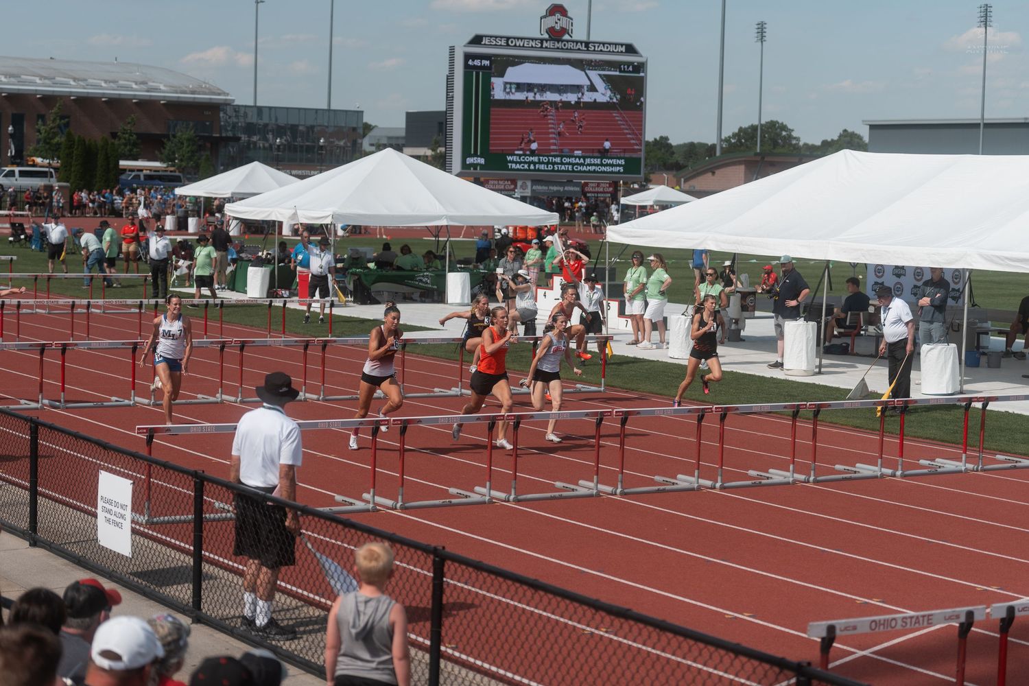 Hurdles race competitors approach finish at outdoor track stadium with scoreboard visible in background.