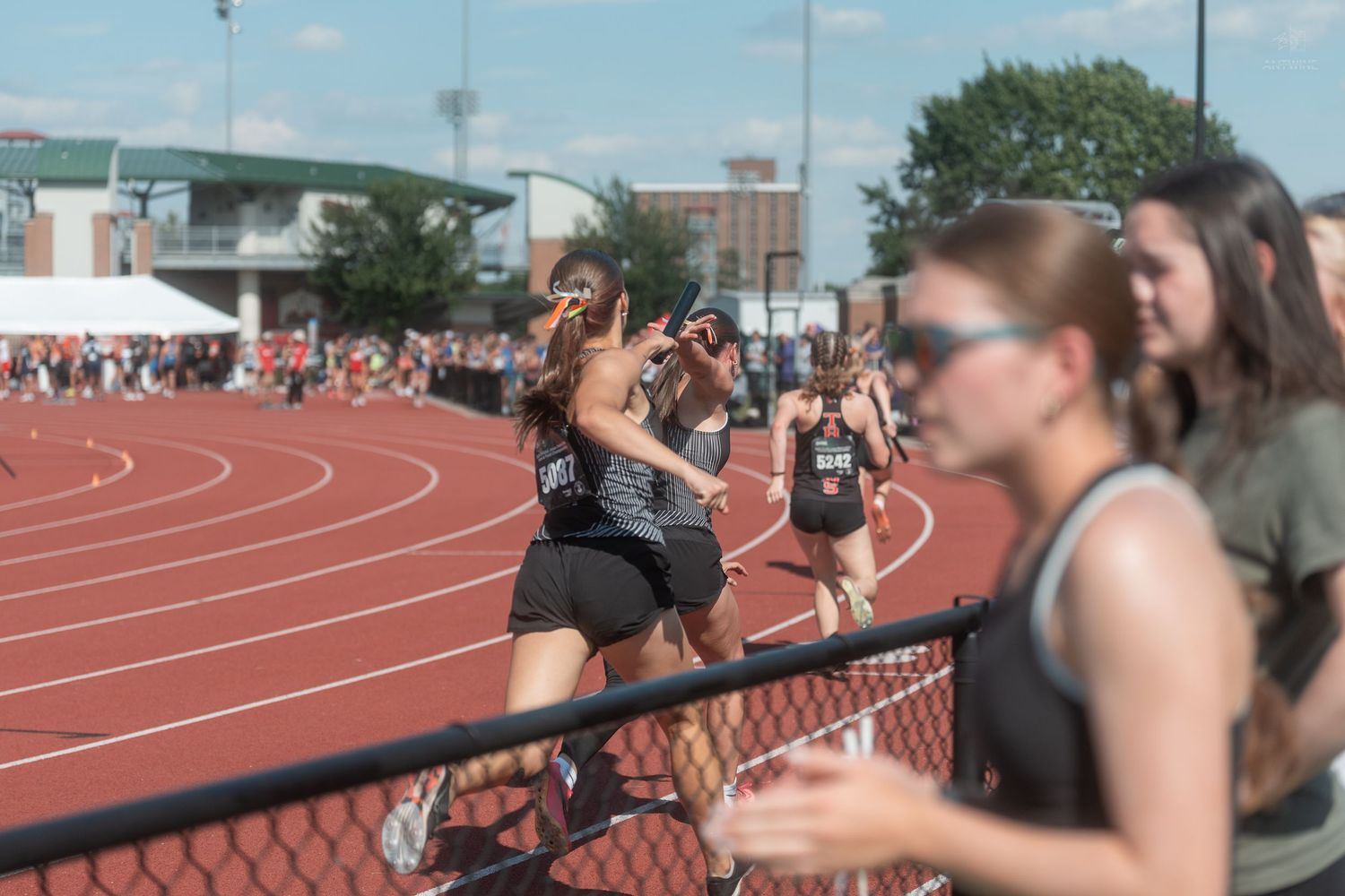 Female runners compete on a red track during an outdoor track and field meet on a sunny day.