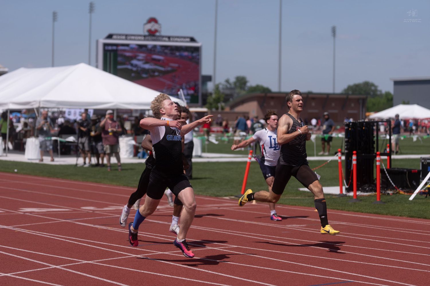 Male athletes compete in a sprint race at an outdoor track and field event on a sunny day.