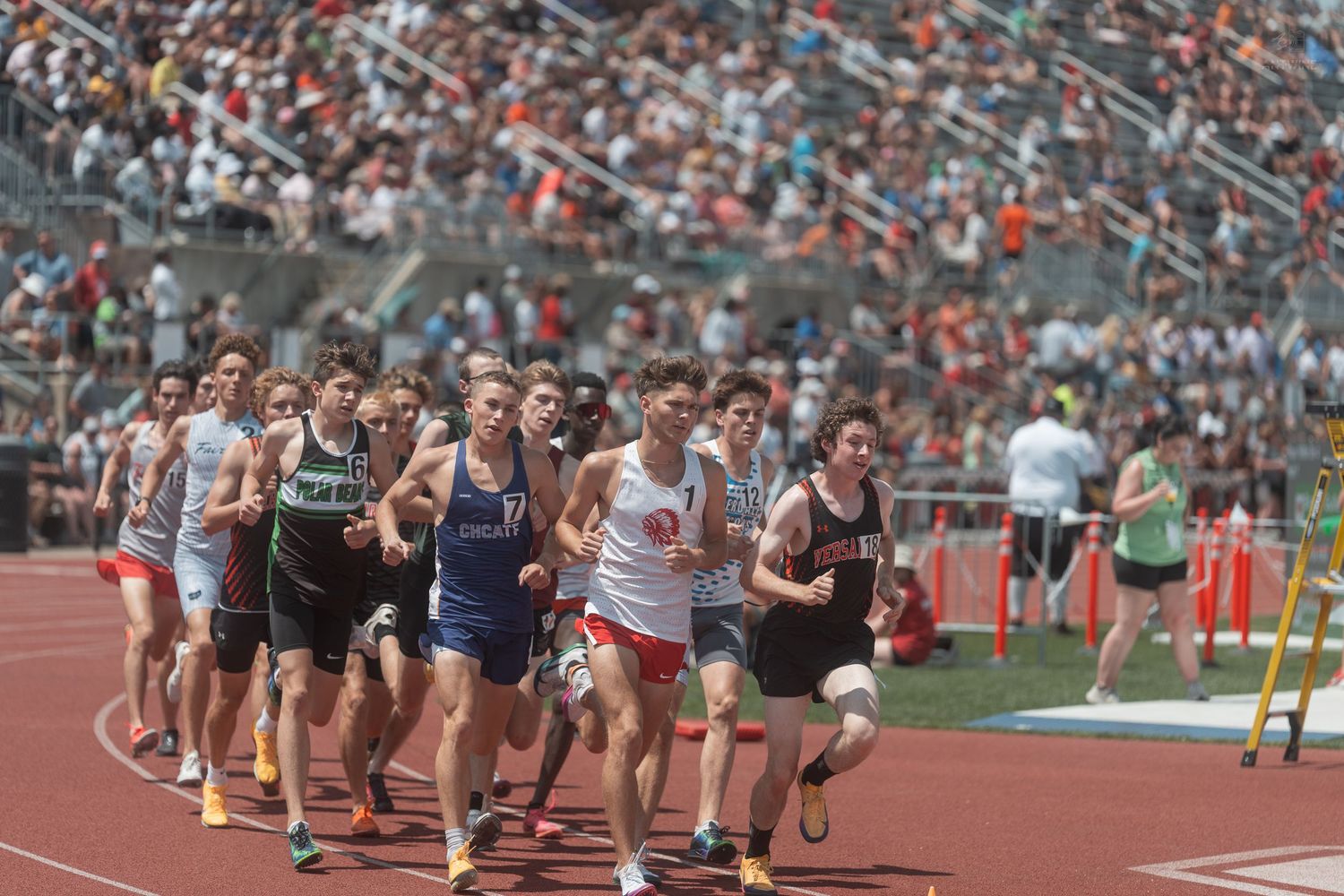 Large pack of high school runners compete in a distance race on a red track at a crowded state track meet.