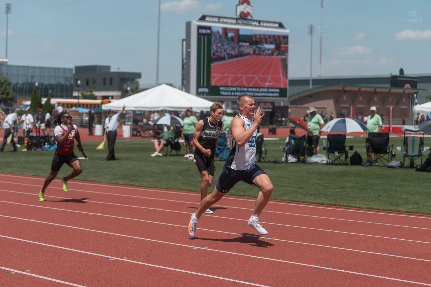 A sprinter races down the track in focus while competitors follow behind at an outdoor track and field event.