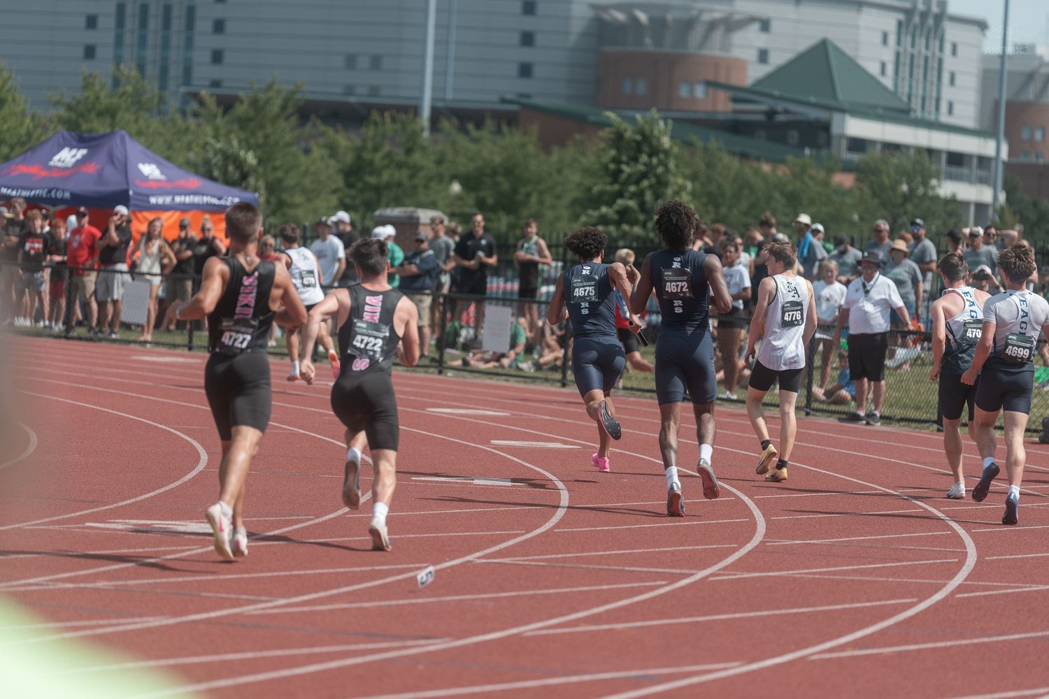 Athletes compete in a relay race on a red track at an outdoor high school track and field meet.