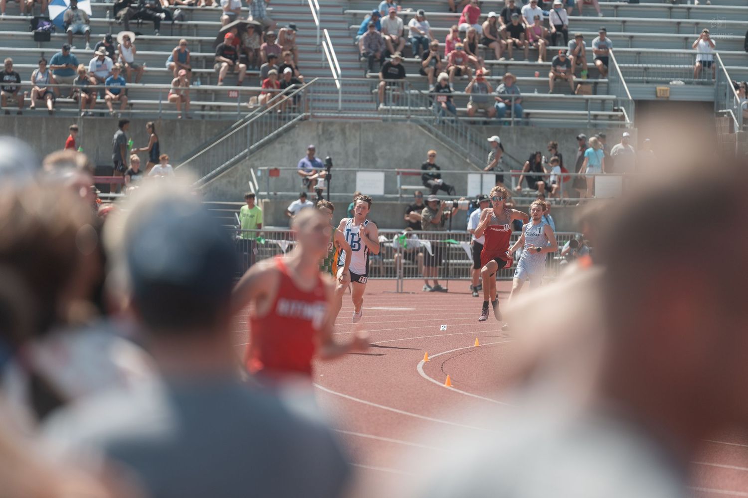 Crowd watches male runners compete on red track during outdoor high school track and field competition.