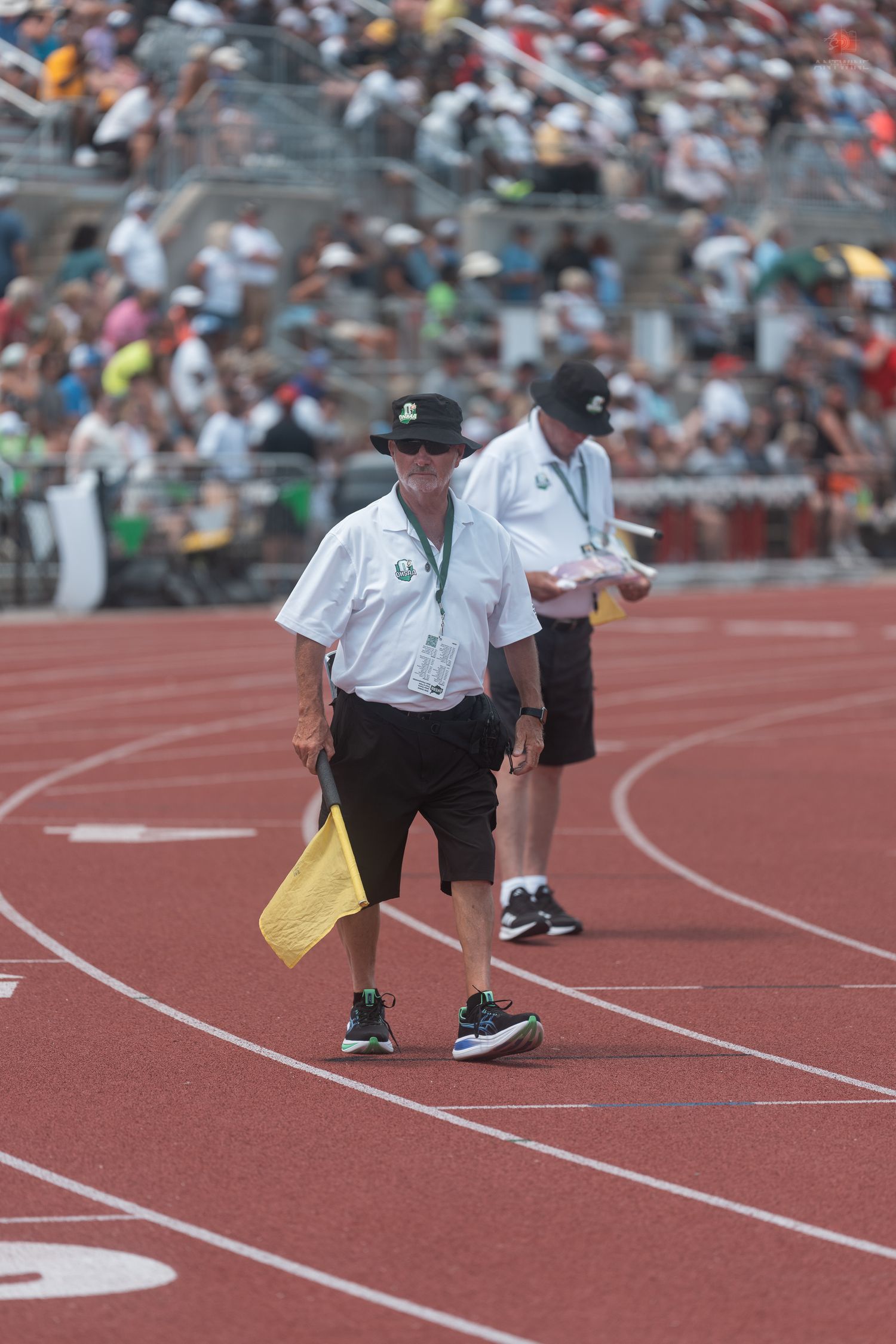 Track official in white shirt carries a yellow flag on a red running track at a crowded outdoor athletics meet.