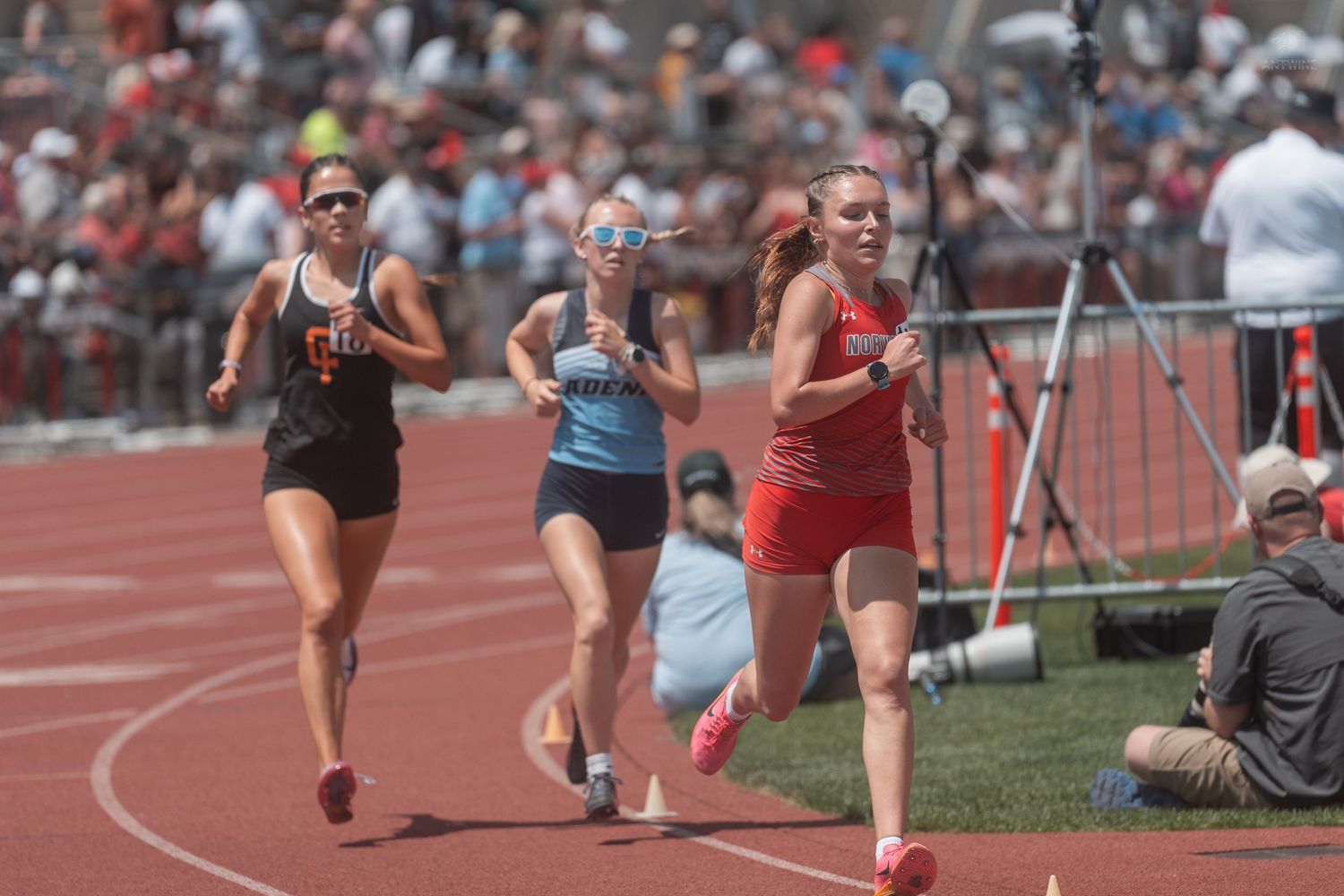 Three female athletes compete in a track race on a red oval during a sunny outdoor high school track and field meet.