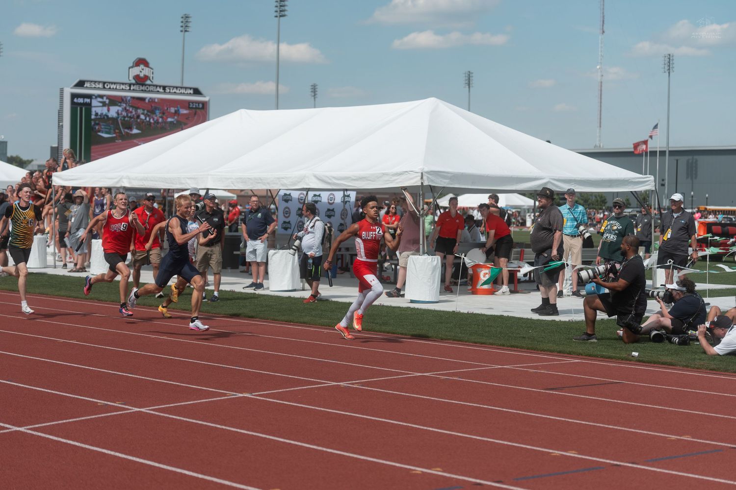 Multiple athletes race on red track at outdoor track and field competition under bright sunny skies.