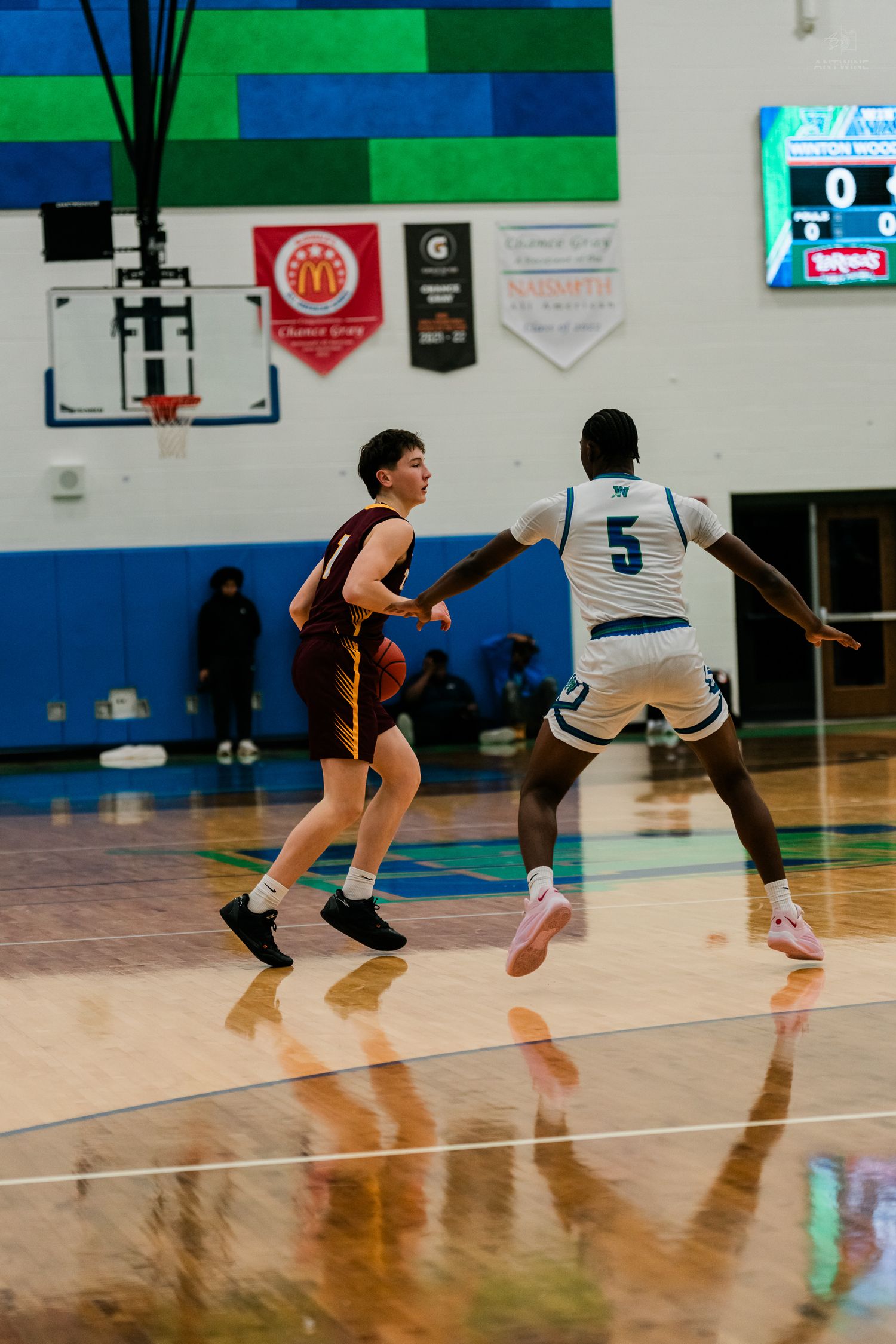 Player #5 in white defends against a maroon-uniformed opponent dribbling on a shiny hardwood basketball court.