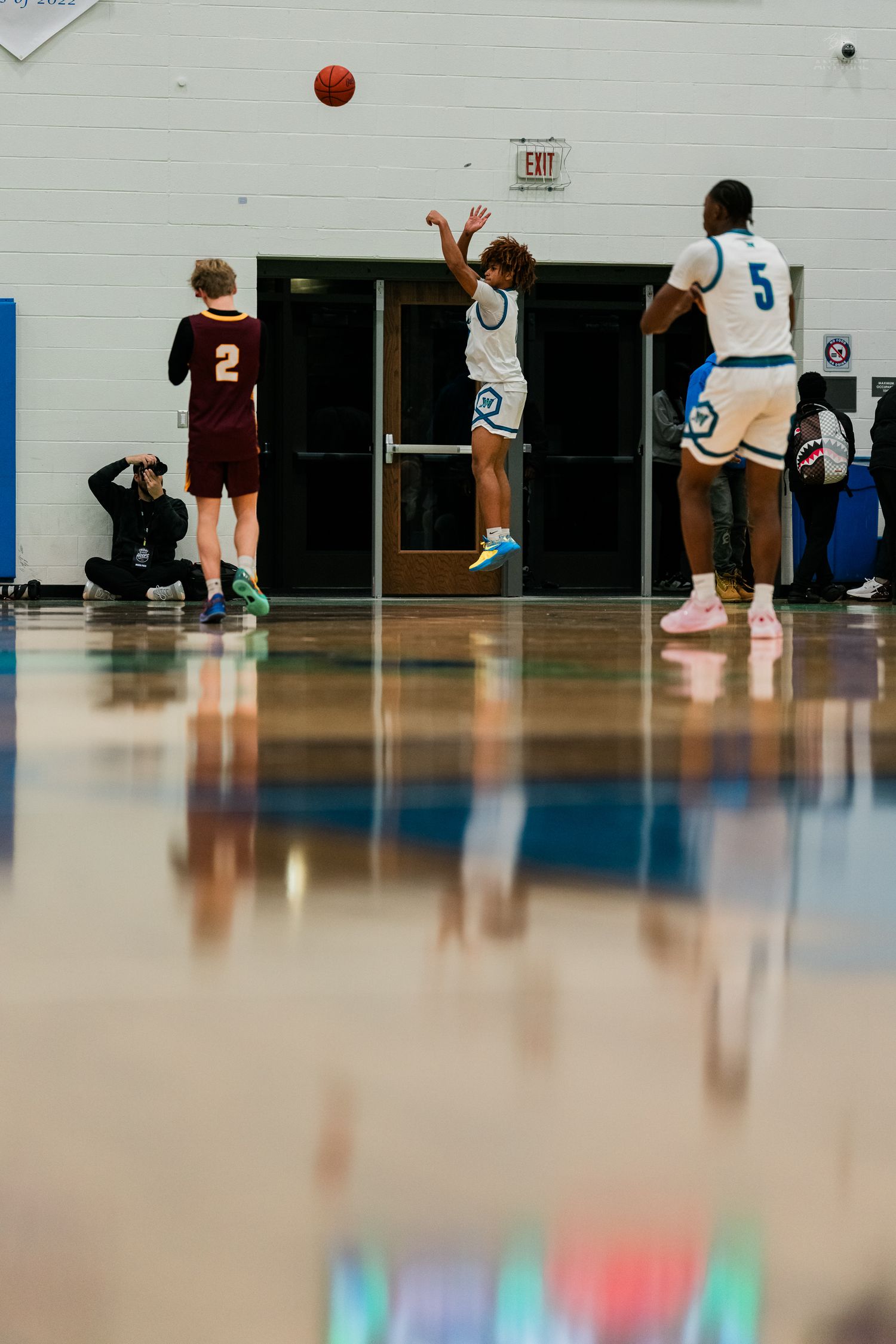 High school basketball player in white uniform jumps to shoot while teammates and opponent watch during indoor game.