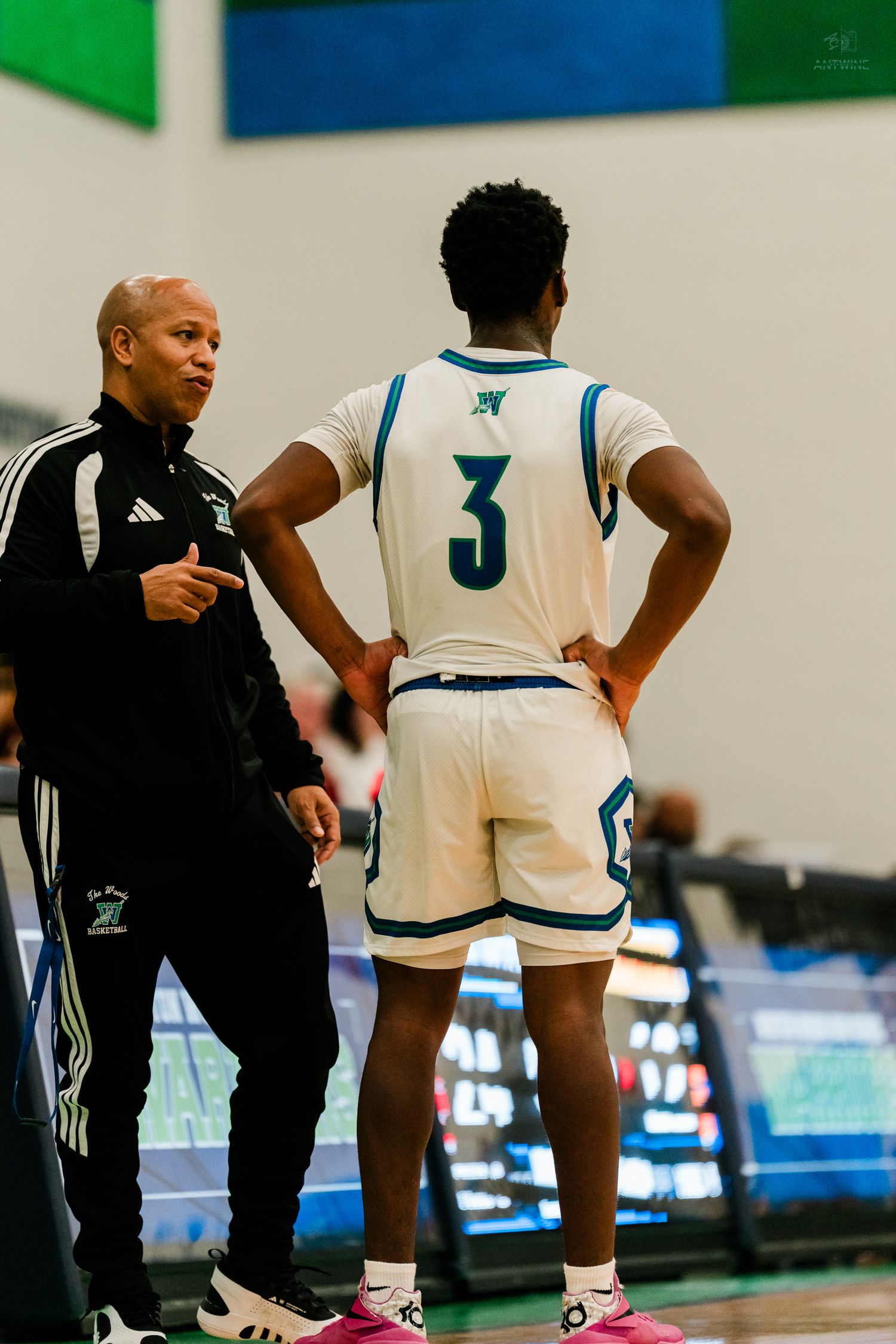 Basketball coach in black Adidas tracksuit talks to player #3 in white and teal uniform during game.