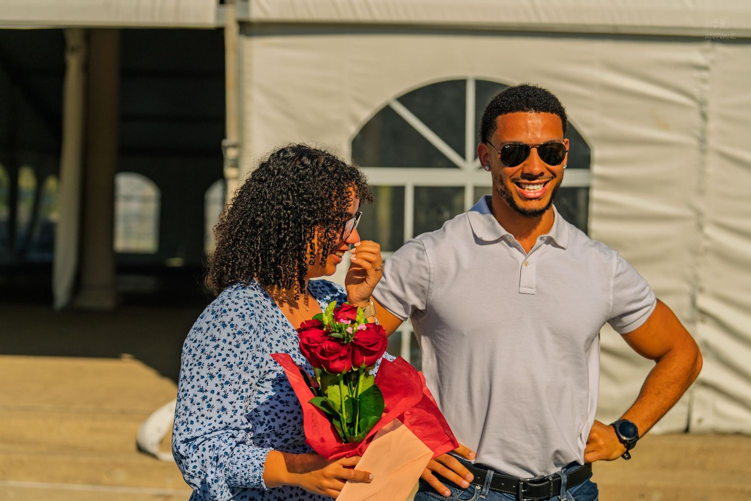A joyful couple shares a moment as a woman smells a bouquet of red roses gifted by a man in sunglasses.