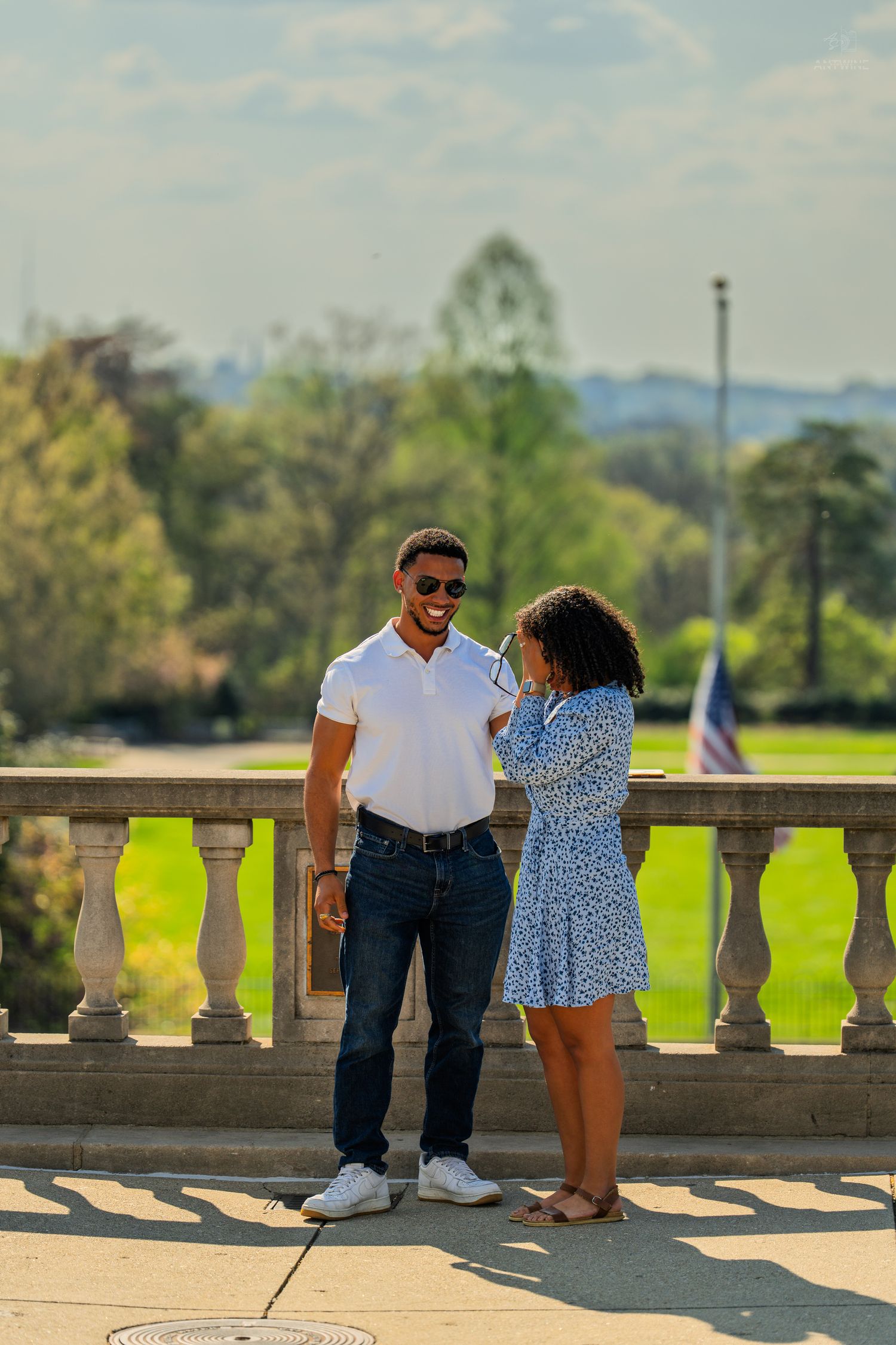 A couple smiles at each other on a stone balcony terrace with lush green park views behind them.