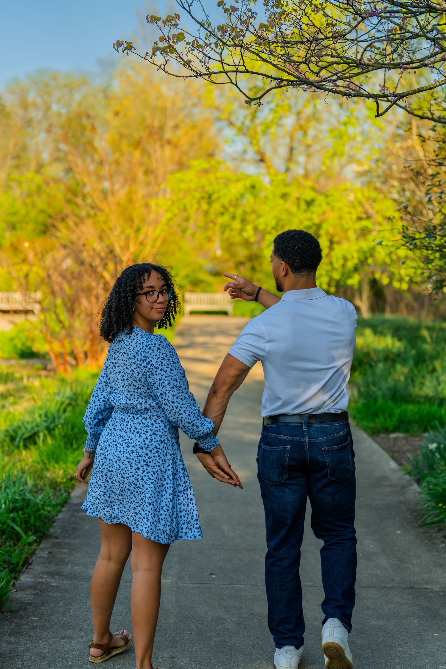 Couple walking hand in hand on a sunny park path, woman in blue dress looking back smiling.