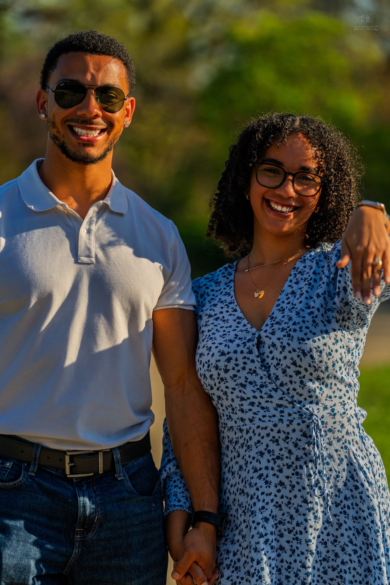 Smiling couple holding hands outdoors in golden sunlight, man in white polo and woman in floral dress.