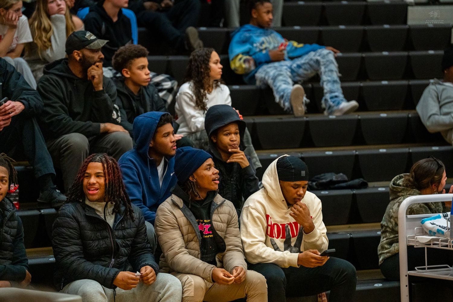 Young spectators sitting in gymnasium bleachers watching a high school basketball game intently.