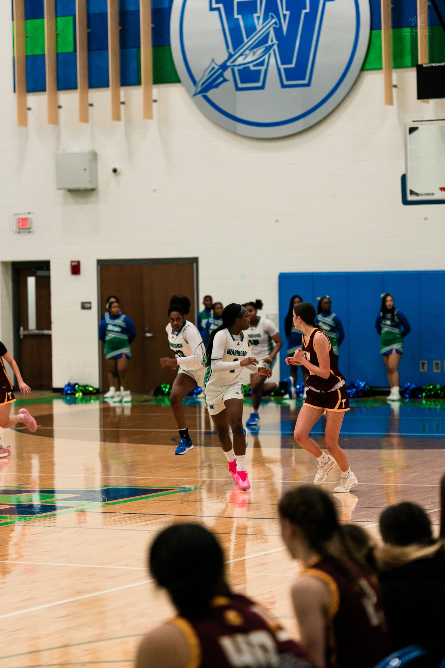 Female basketball player in white uniform drives past defender in maroon jersey beneath a large W logo in gymnasium.