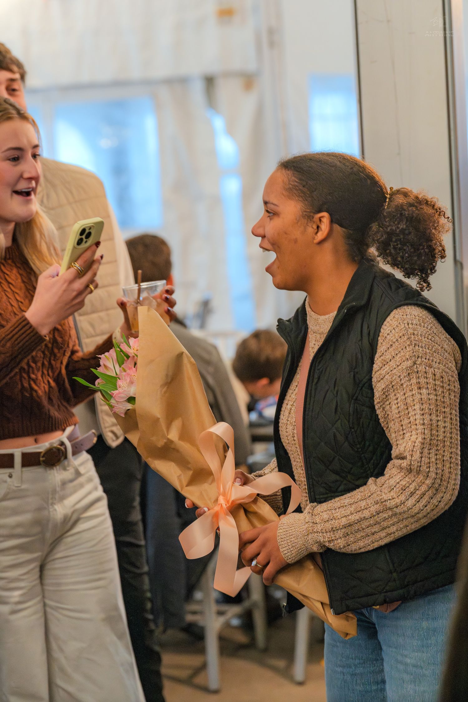 A joyful young woman receives a bouquet of pink flowers wrapped in kraft paper at a celebration.