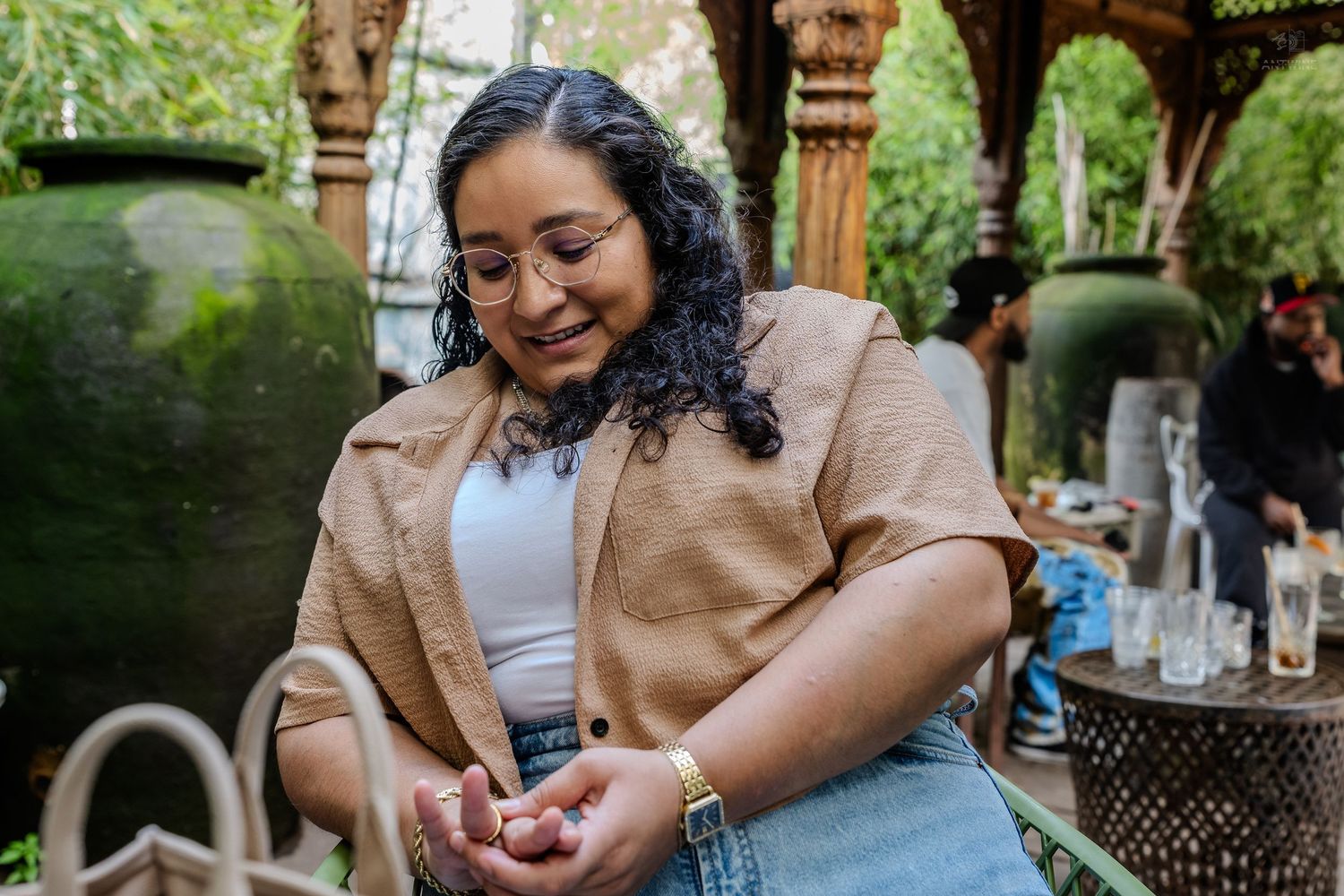 Young woman in tan blazer and jeans smiling while looking at her hands in a garden setting.