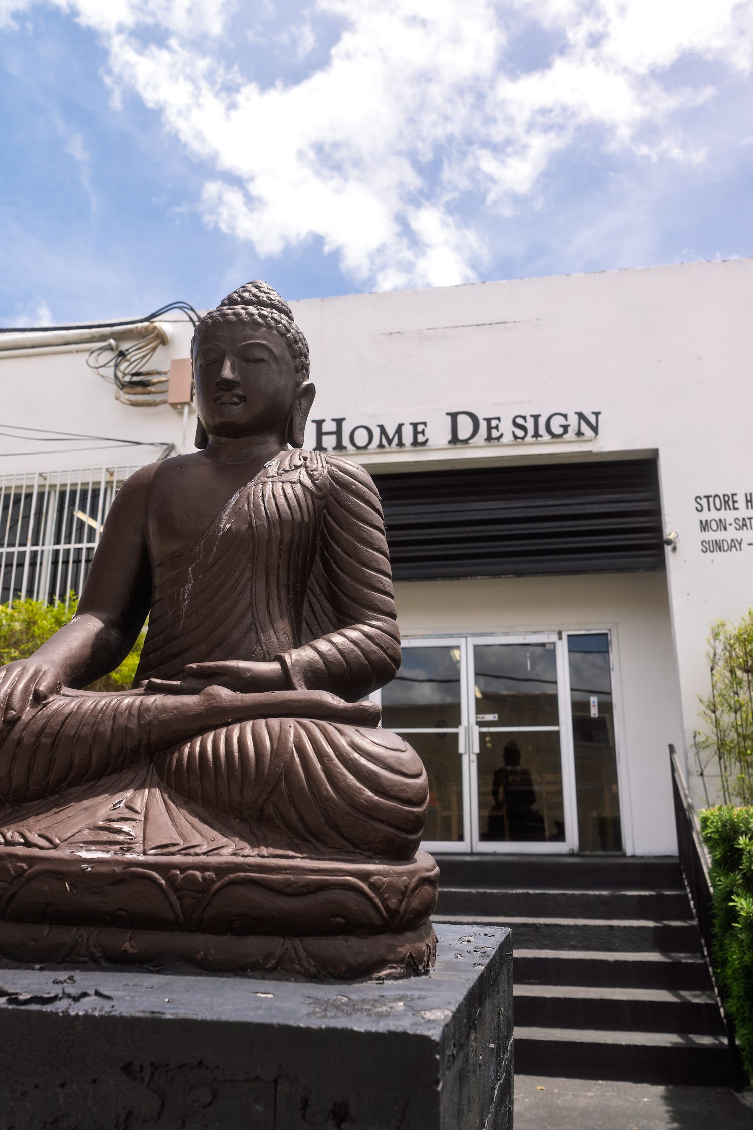 A large bronze Buddha statue sits in meditation outside a Home Design store under blue skies.