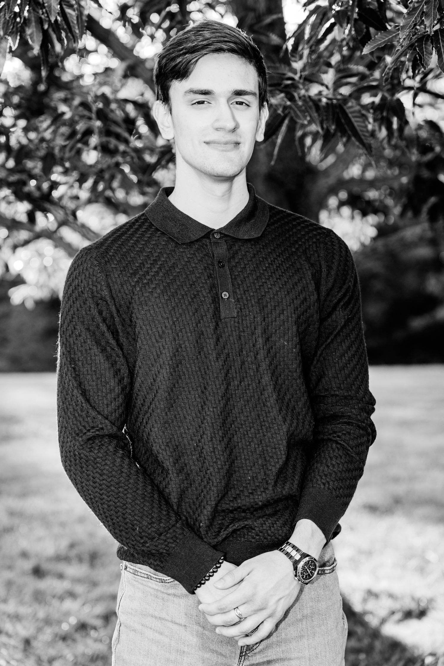 Young man in black polo sweater smiling outdoors with trees in background, black and white portrait photo.
