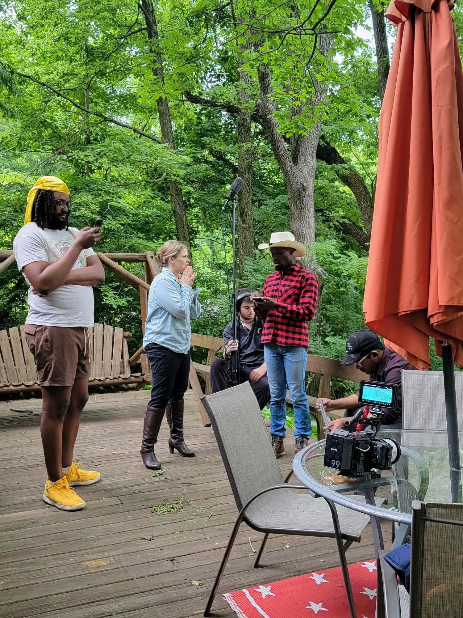 Film crew on wooden deck with red umbrella, surrounded by lush green trees, filming outdoor scene.