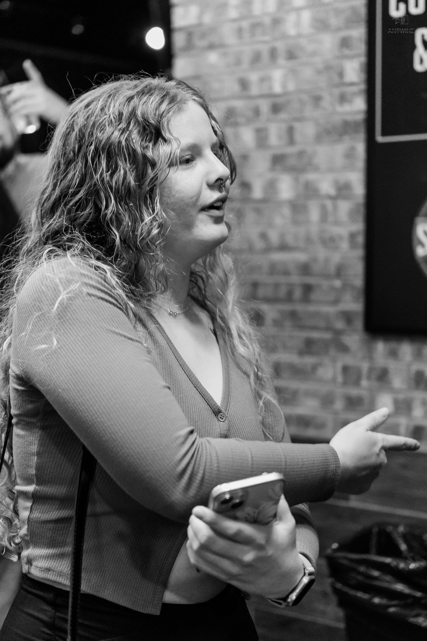 Black and white photo of a curly-haired woman holding a smartphone and gesturing while talking at a bar.